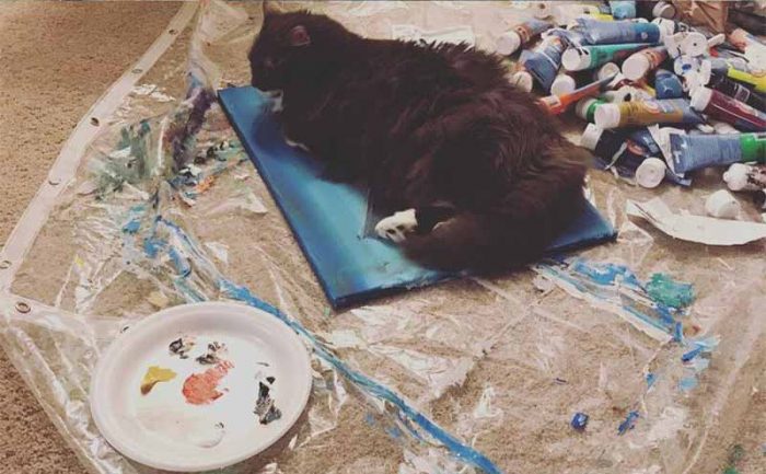 A cat lying on a painting which is next to paint tubes and a plate with small amounts of paint on it 
