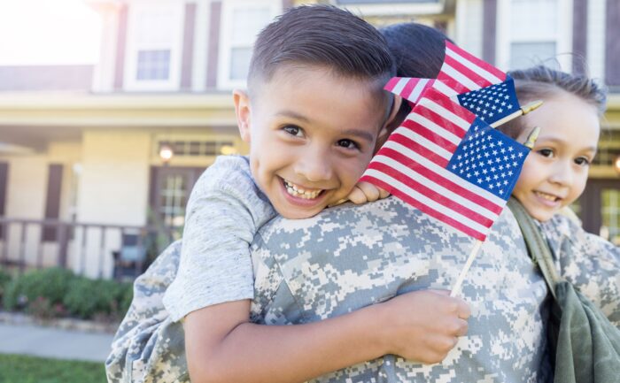 A young boy smiles as he holds flags and hugs his mother in uniform.