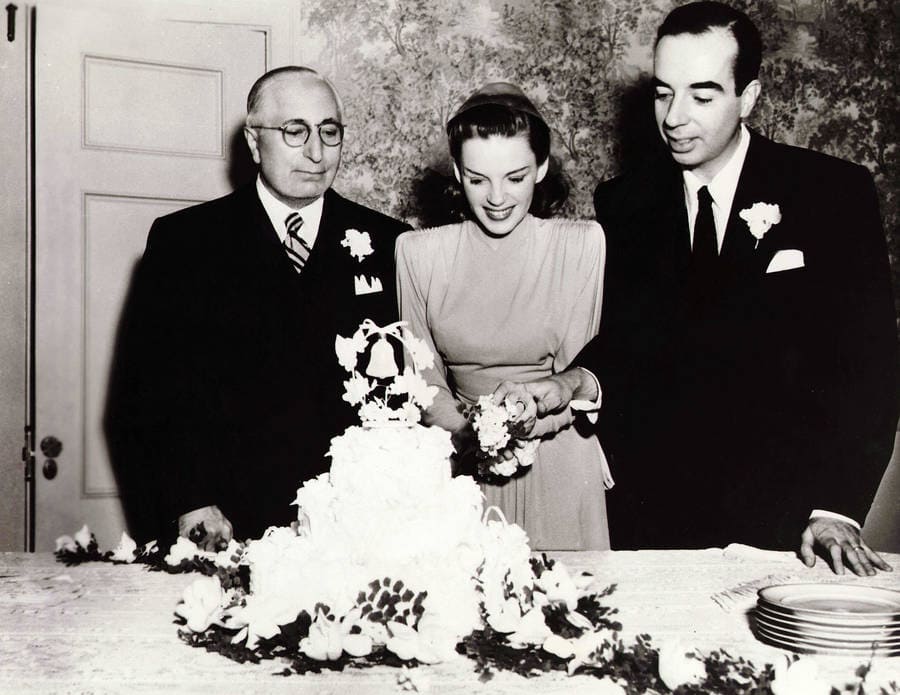 Judy Garland and Vincente Minnelli cutting a wedding cake together while Louis B. Mayer stands by.