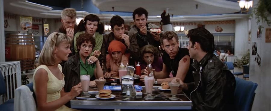 A shot of Olivia Newton-John and John Travolta sitting in a diner drinking shakes with their friends surrounding them. 