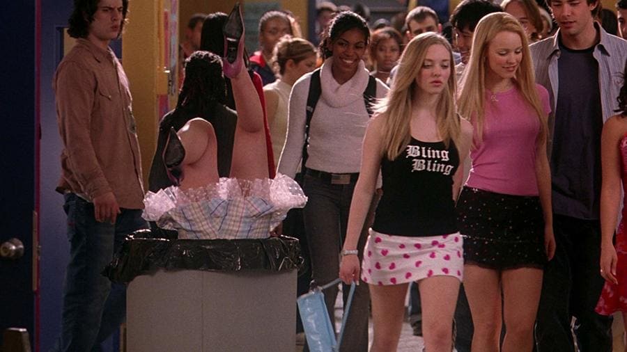 Scene with Rachel McAdams, Jonathan Bennett, and Amanda Seyfried walking away smiling as Linsay Lohan’s feet stick out of the garbage bin in a high school setting. 