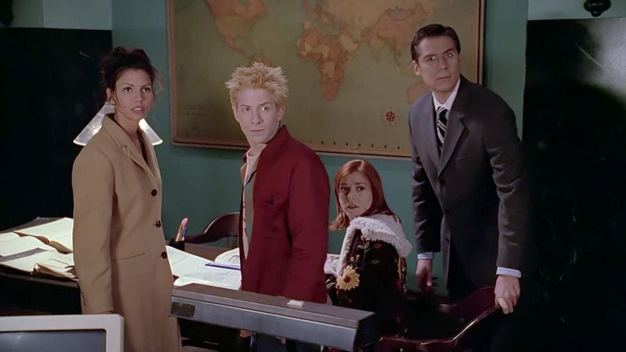 Seth Green, Charisma Carpenter (left), Alyson Hannigan, and Alexis Denisof are hanging around a desk in Buffy the Vampire Slayer. 