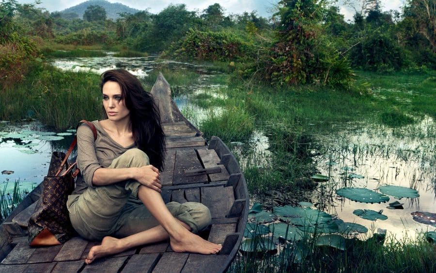 Angelina Jolie on a green river in a wooden boat with lilypads and grass everywhere, holding a Louis Vuitton bag.