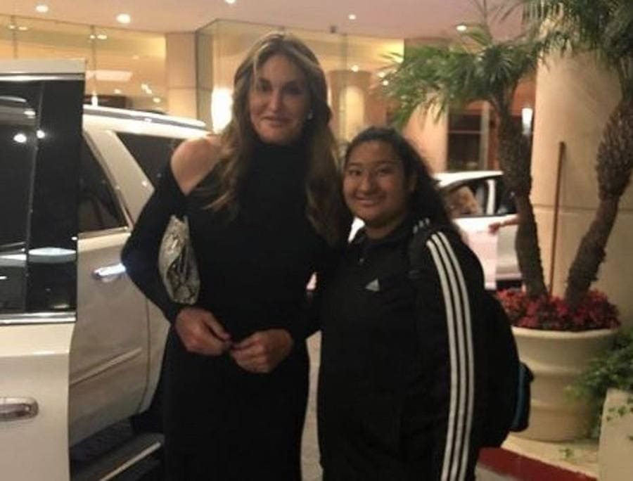 Caitlyn Jenner is posing with a long black dress on with one shoulder out and with Marlen next to a large white car.