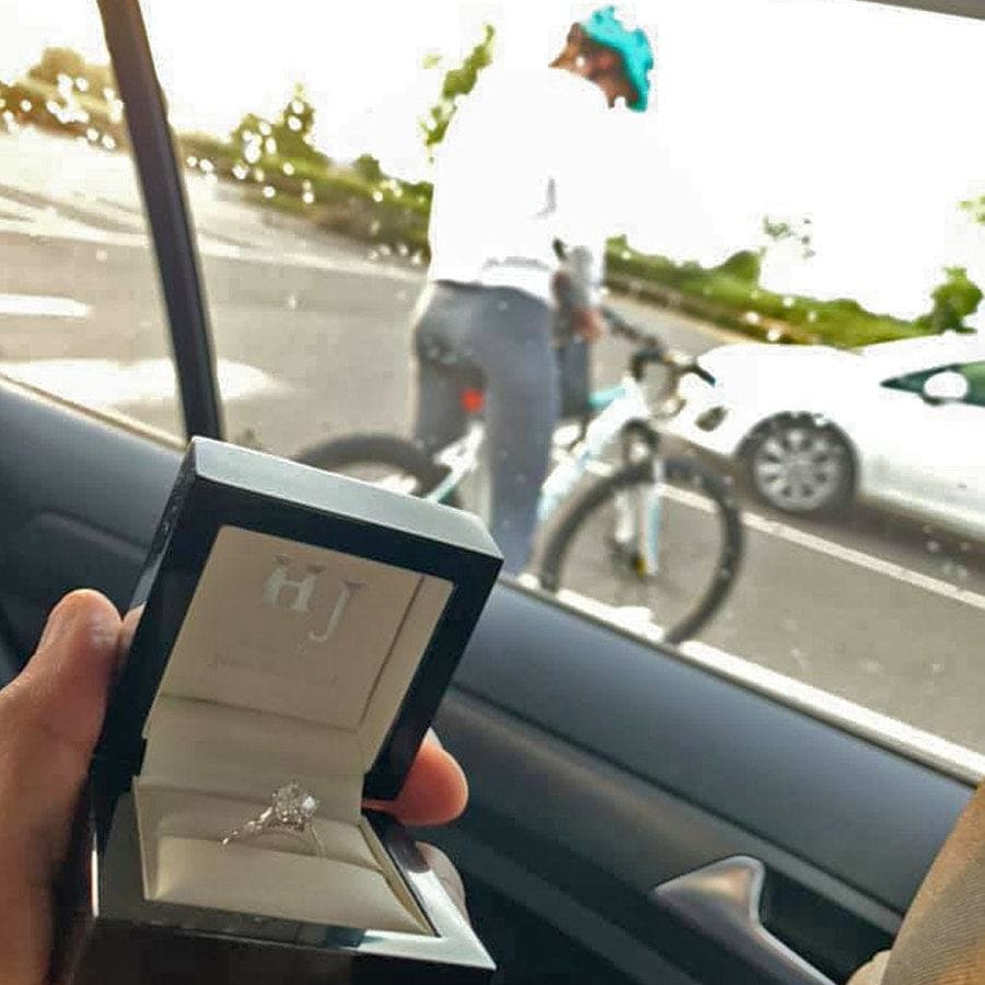 Cally on her bike outside of the car window while Edi holds up the engagement ring box behind her back.