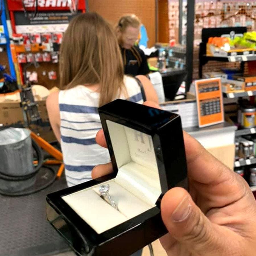 Cally is paying the cashier at a bike shop while Edi proposes behind her back.