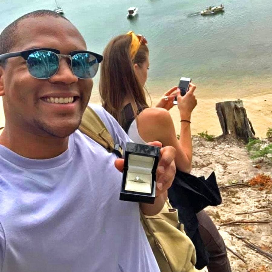 Edi is taking a selfie of the engagement ring behind Cally’s back while she’s on her phone next to the ocean.
