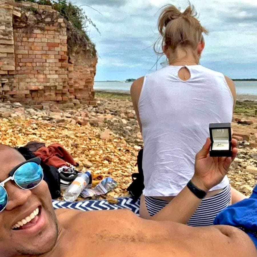 Edi is taking a selfie with the ring behind Cally’s back while she sits on her phone in front of the ocean.