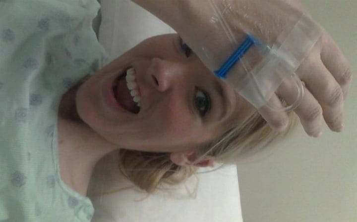 Mandi with an IV in her arm, sitting in a hospital bed with a hospital gown on.