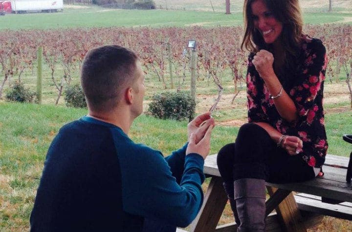 Tyler on one knee proposing to Mandi, who is sitting on a picnic table.