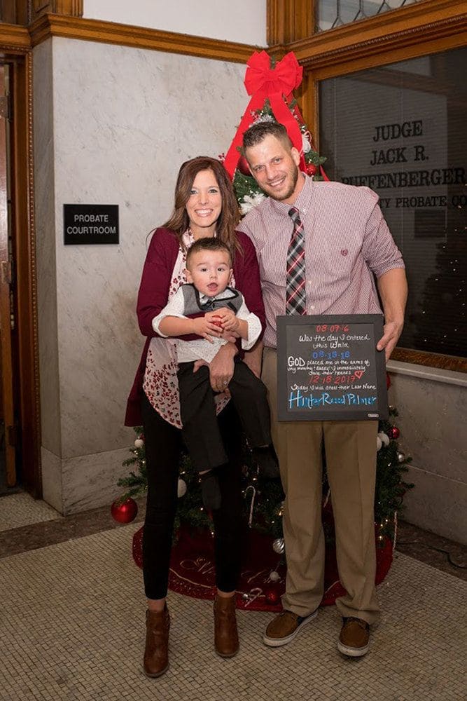 Mandi, Tyler, and Hunter in front of a Christmas tree in the courthouse, with Tyler holding a board with the adoption information written.