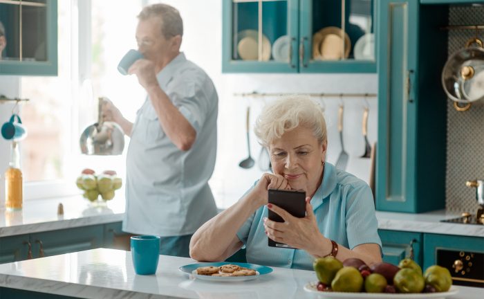 An older woman on the phone with her husband drinking a cup of tea behind her in the kitchen