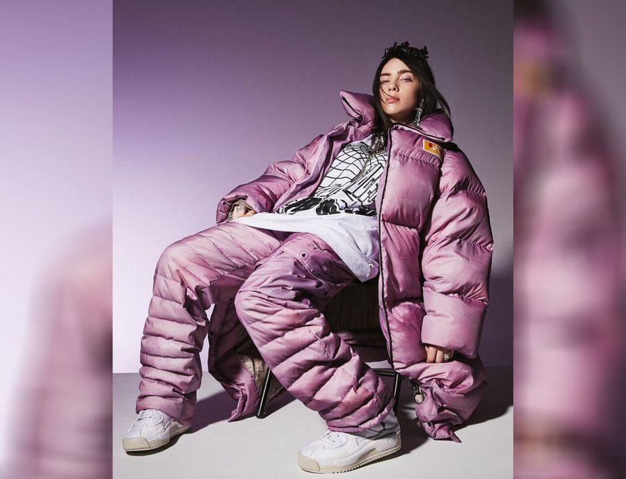 Billie Eilish wearing a large light purple puffy jacket and puffy pants with white sneakers and a white t-shirt. 
