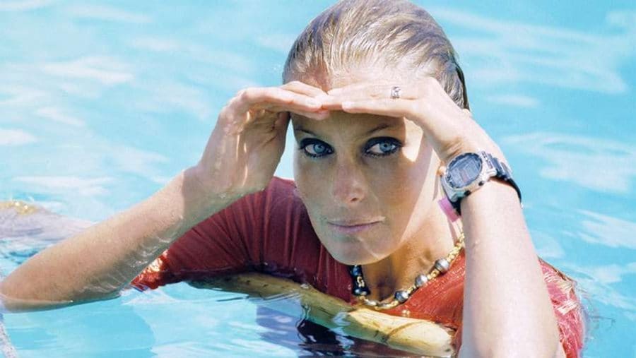 Bo Derek is sitting in the swimming pool with an orange shirt on and her hair wet. 
