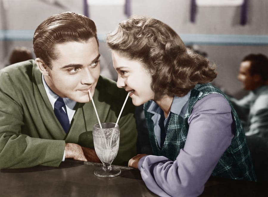 A couple in a restaurant looking at each other and sharing a milkshake with two straws