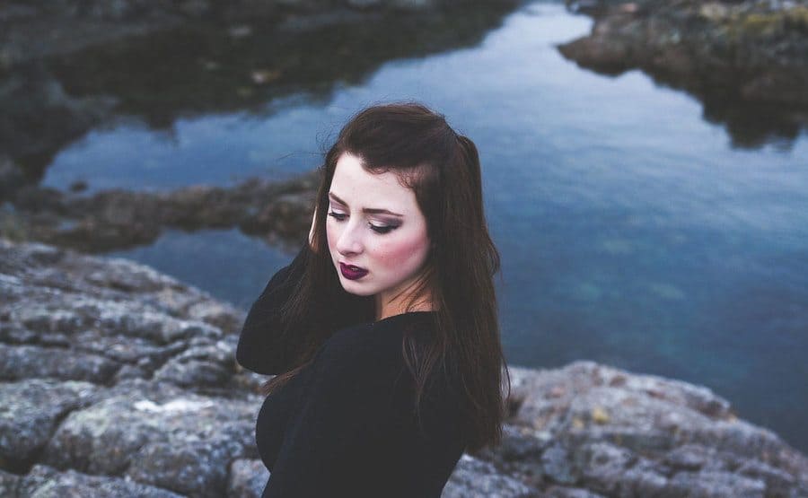 Laura in dark clothing with brown hair and dark red lipstick photographed in front of the water with large rocks around 
