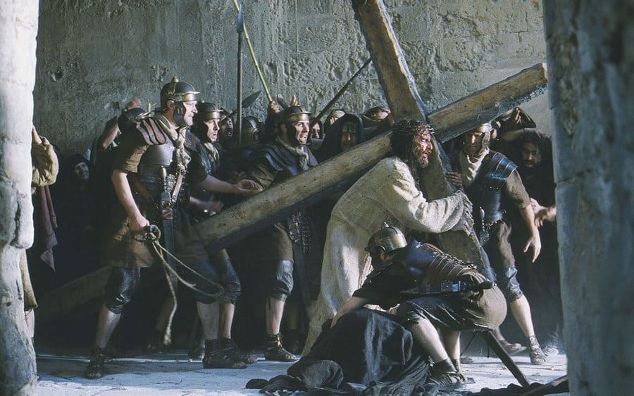 Jim Caviezel pulling a large cross on his back while guards whip him in ‘The Passion of the Christ.’