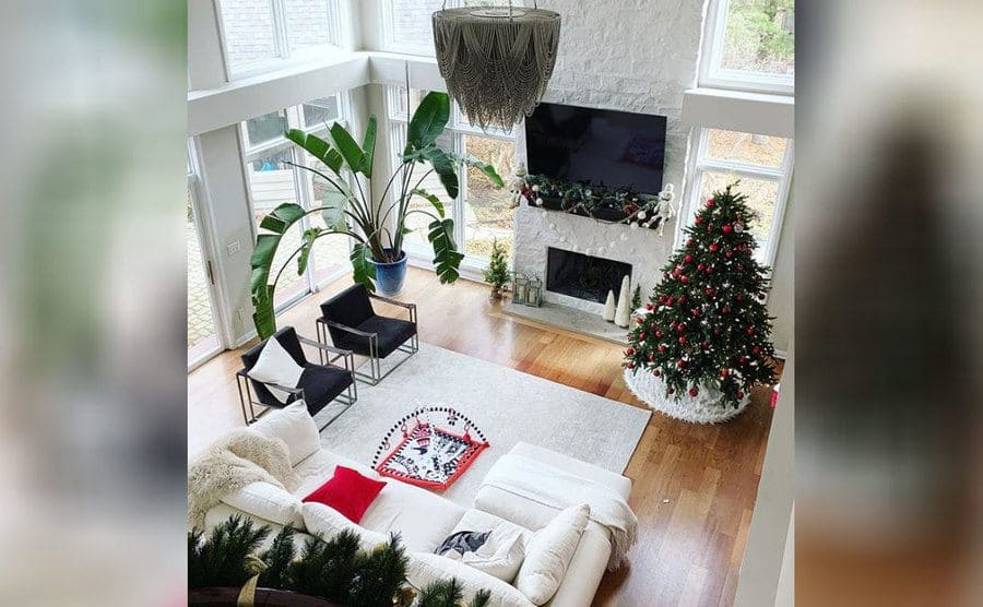 A photograph of Myka’s white living room with a tall ceiling and wooden floors, a lot of windows, a large TV, an intricate silver chandelier, and Christmas decorations