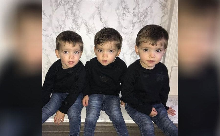 The triplets sitting on a marble ledge in dark jeans and black sweaters 