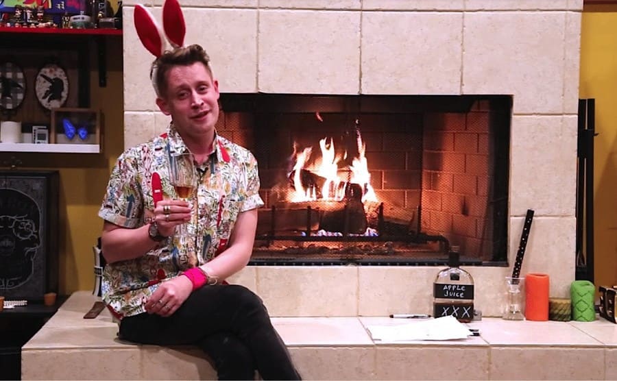 Macaulay Culkin sitting with bunny ears on near a fireplace 