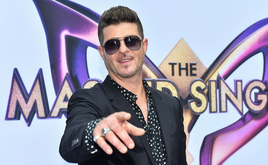 Robin Thicke pointing in front of a sign for the show “The Masked Singer.”