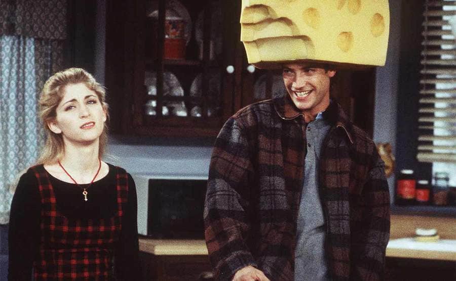 Staci Keanan with Sasha Mitchell standing in the kitchen while Sasha wears a large piece of cheese on his head in the show Step by Step 