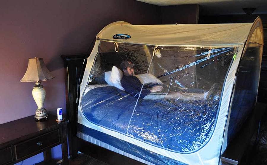 A man lying on a bed with his laptop inside of a hypoxic tent