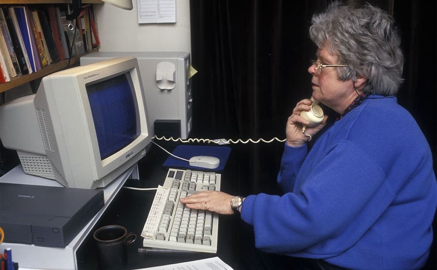 An older woman using an old computer to look stuff up while on the phone 