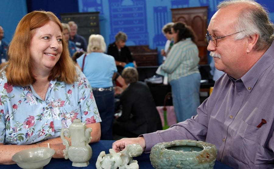 A woman with jade bowls and other decorative pieces talking to an appraiser 