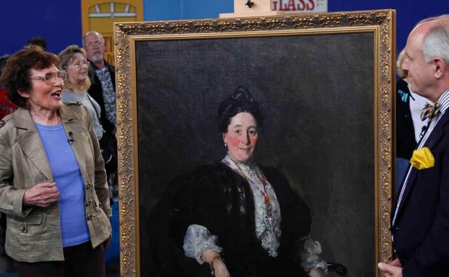 An older woman excited while an appraiser looks at a large older painting 