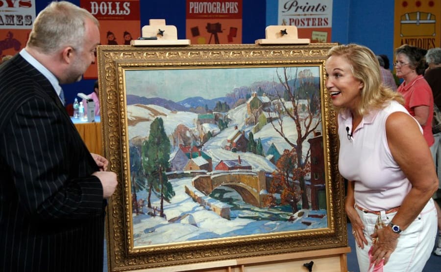 An excited woman with an appraiser standing in front of a painting of a snow-covered village 