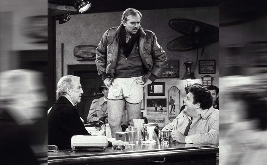 John Ratzenberger standing up on the barstool while Nick Colasanto and George Wendt sit around him 
