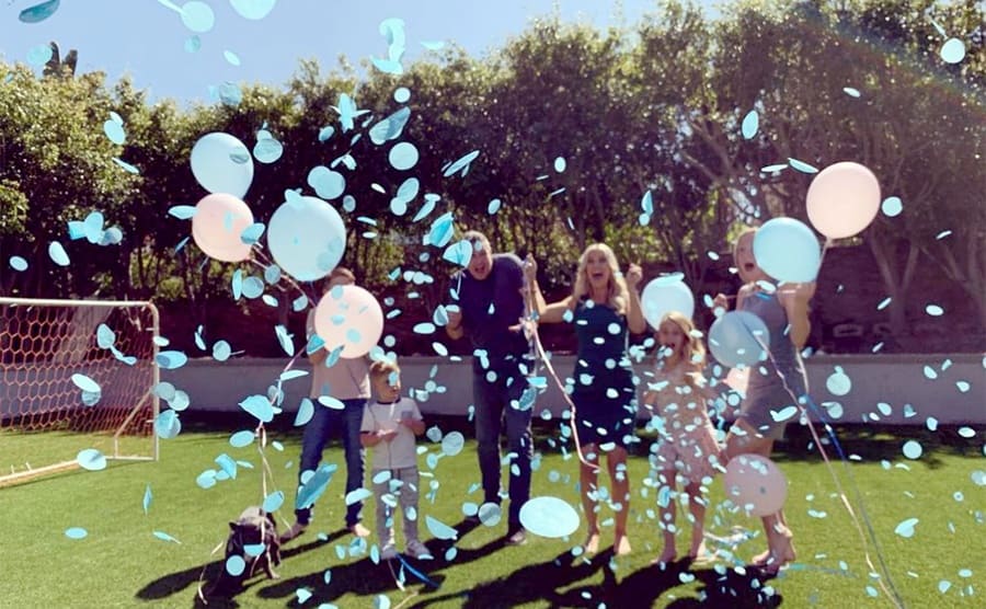 Anthony, Christina, and their children standing on a small soccer field excited with blue and white balloons and confetti in the air 