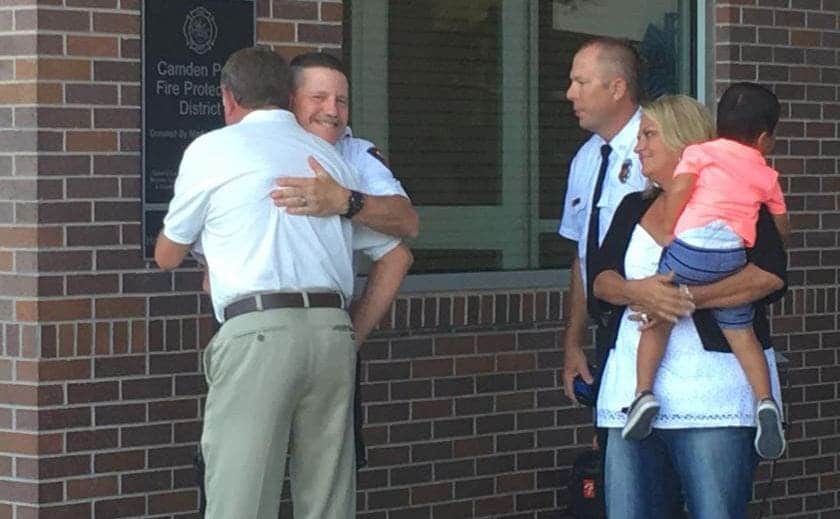 Mark Hill hugging the fire chief while his wife holds their child 