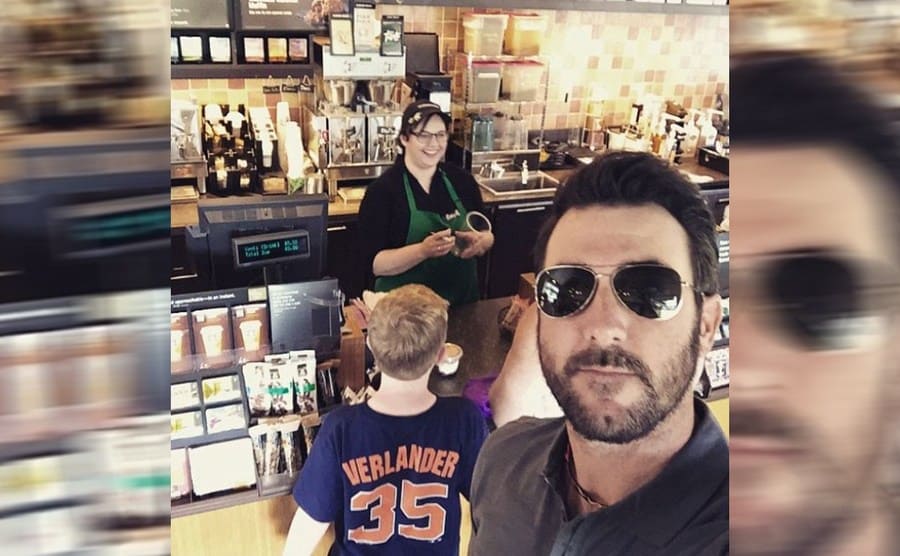Justin Verlander taking a selfie with a kid wearing a baseball shirt with his last name on it while standing in line at Starbucks with his back turned towards the counter