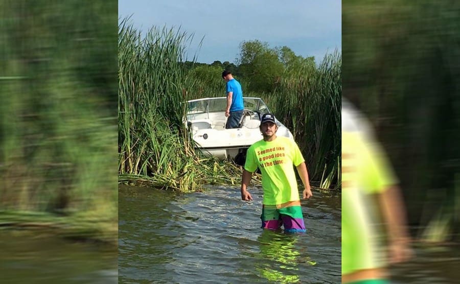 A guy wearing a shirt that says seemed like a good idea at the time while standing in the water with his boat with his friend inside is stuck up in the shrubs 