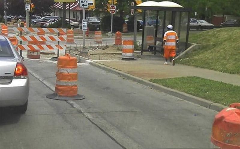 A construction zone with orange and white cones and barriers with a man walking in an outfit that matches the construction zone 