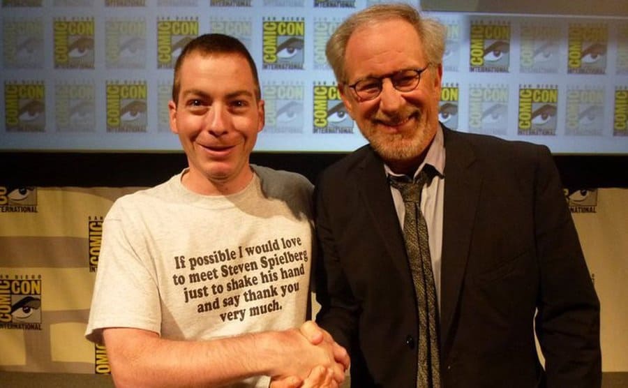 A guy with a t-shirt that says ‘If possible I would love to meet Steven Spielberg just to shake his hand and say thank you very much’ on it shaking Steven Spielberg’s hand 