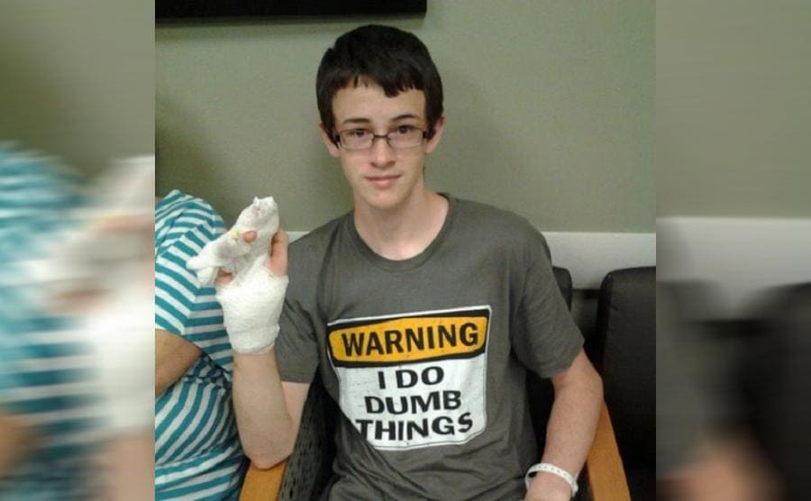 A kid wearing a t-shirt in an ER that says Warning I do dumb things while he holds his bandaged hand up 