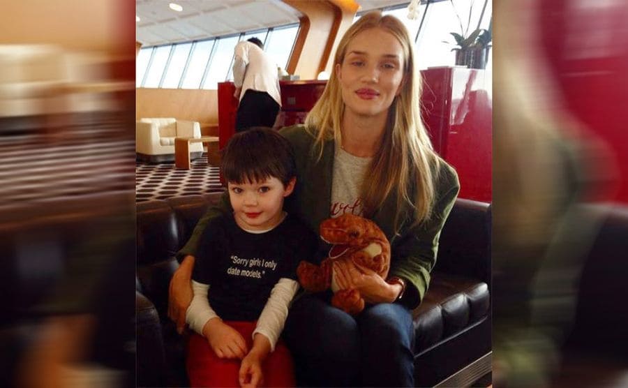 A kid wearing a shirt that says ‘Sorry girls, I only date models’ while posing with Victoria’s Secret model in an airport 