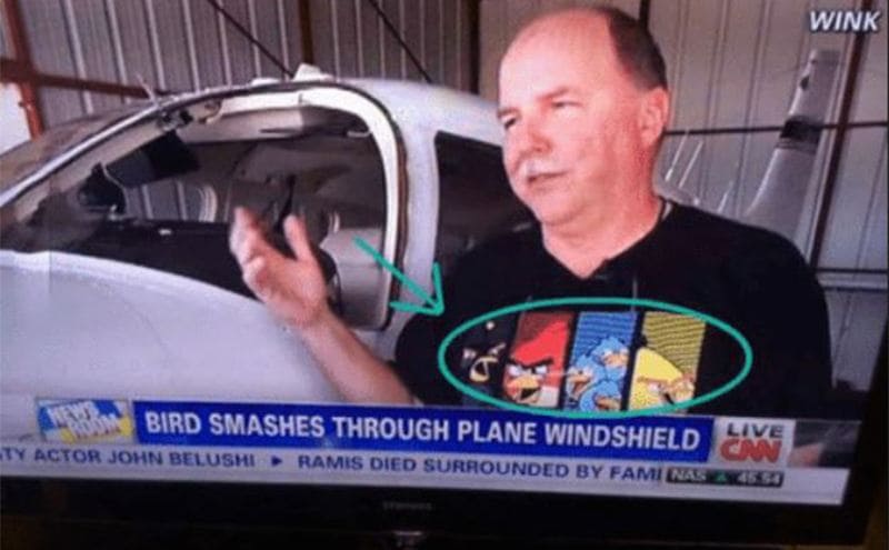 A photograph of the news on a tv screen with a guy being interviewed about a bird smashing through a plane window while he wears an angry bird t-shirt 