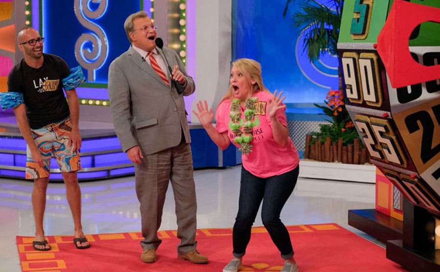 Drew Carey with a microphone standing with a woman who is excited about where she landed on the wheel 