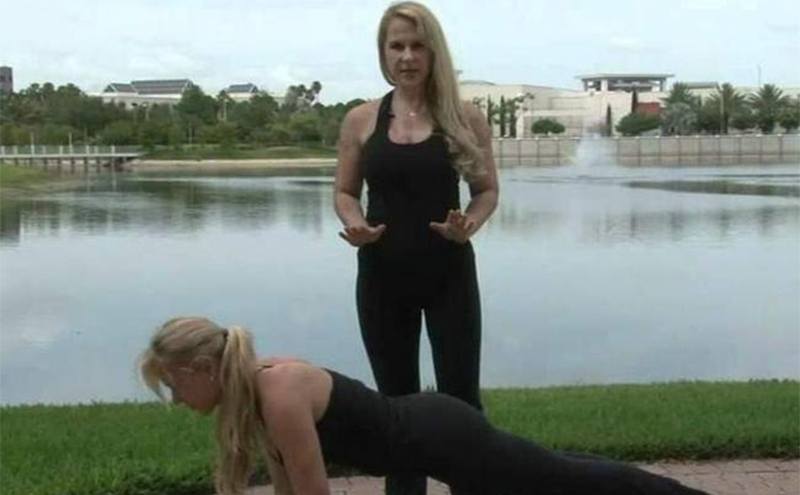 One of the twins doing a yoga pose while the other one stands above her explaining something next to a small lake 