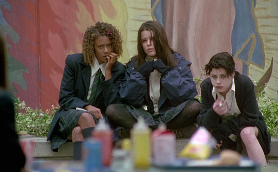 Rachel True, Neve Campbell, and Fairuza Balk sitting on a bench 