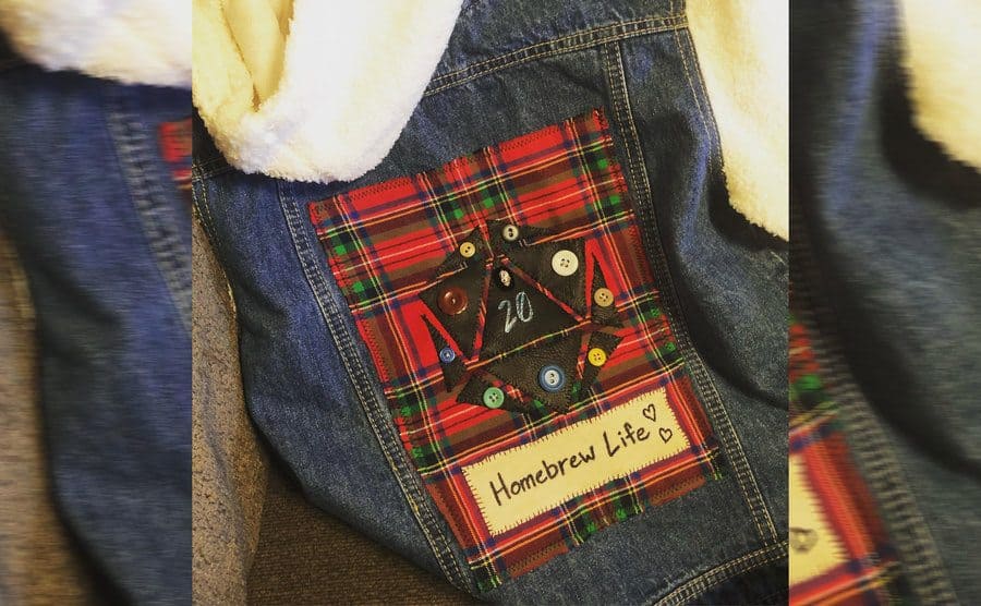 A photograph of the corner of a denim jacket with a red, green, and black plaid patch, black leather with buttons in a symbol, and a patch that says ‘Homebrew Life.’ 