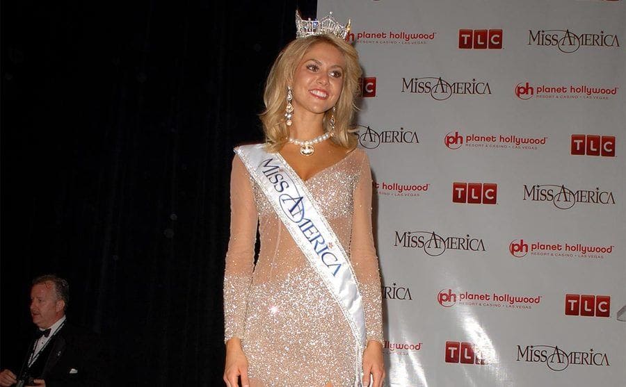 Miss America Kirsten Haglund posing in front of a backdrop with ‘TLC,’ ‘Miss America,’ and ‘Planet Hollywood’ written all over it 