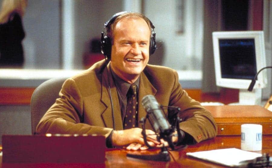 Kelsey Grammar sitting behind a microphone at a desk in a radio station 