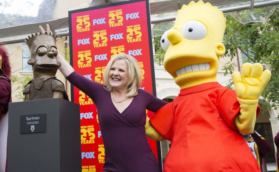 Nancy Cartwright standing in between a man wearing a Bart Simpson costume and a sculptured Bart Simpson head 