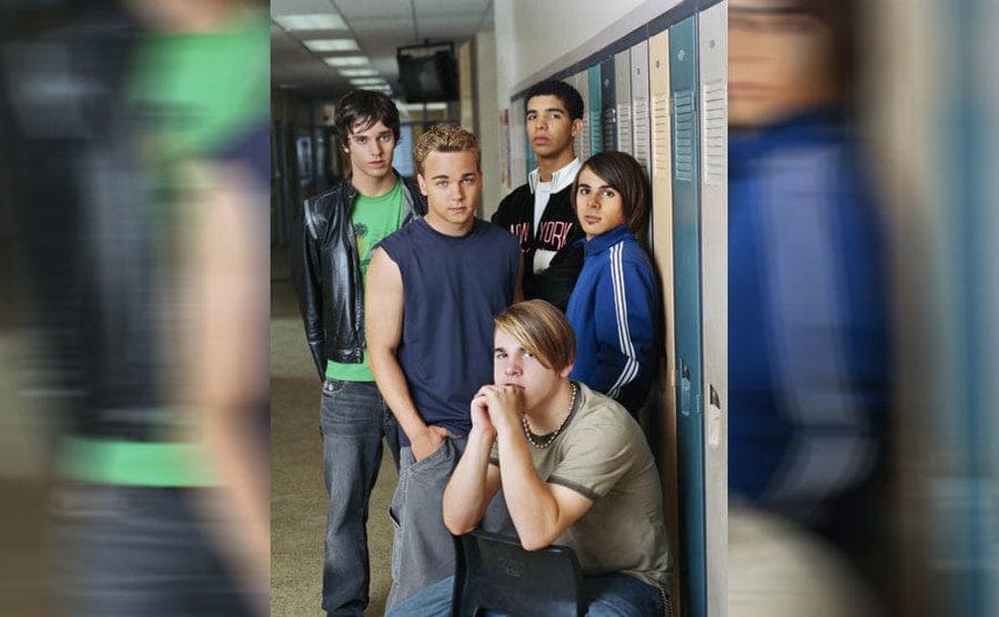 Some of the cast of Degrassi posing with Drake next to lockers in a high school hallway 
