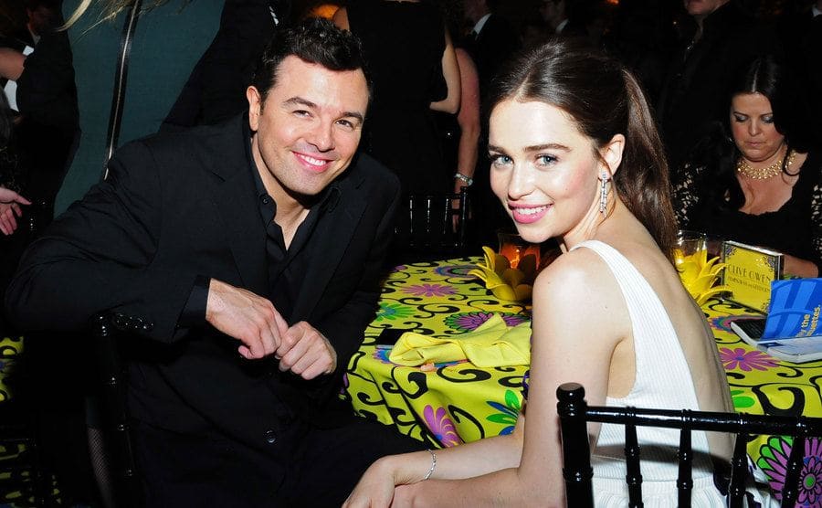 Seth MacFarlane and Emilia Clarke sitting at a table posing for the camera at a party 